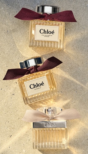 Tři elegantní parfémové lahvičky Chloé s barevnými mašlemi na písčitém pozadí – luxusní vůně s vroubkovaným sklem a kovovým víčkem